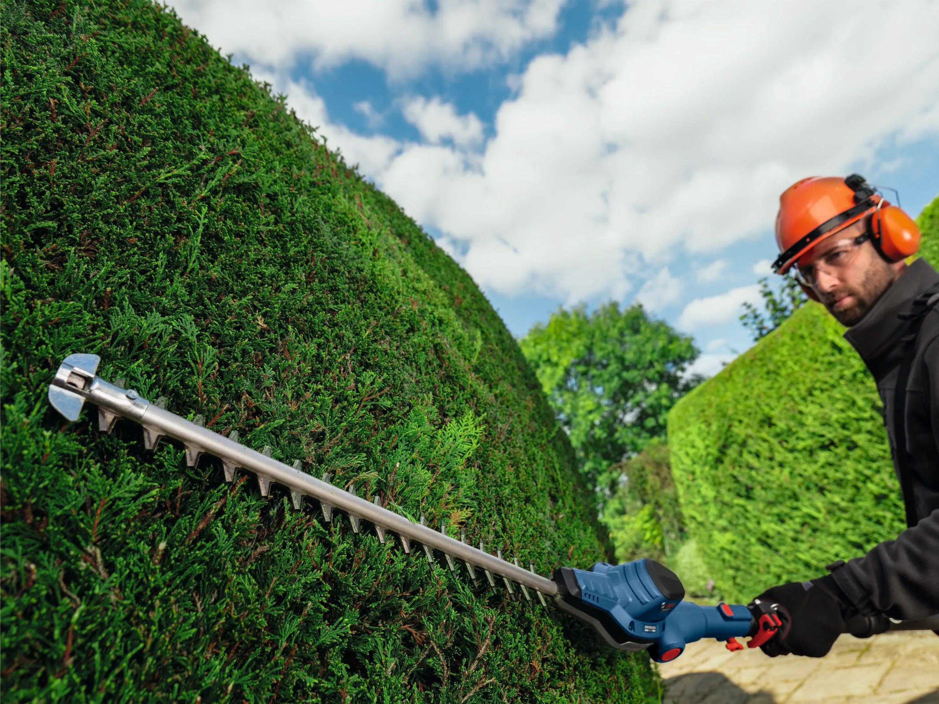 A close-up view of a landscaping contractor wearing an orange hard hat as they guide the 20-inch diamond-ground blade of the Bosch cordless pole trimmer across a tall hedge row under a blue sky with scattered clouds.
