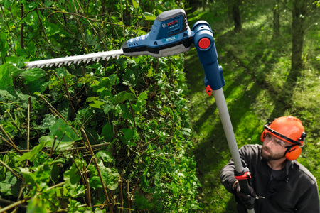 An outdoor view of a property maintenance professional extending the aluminum telescoping shaft of the Bosch 18V brushless hedge trimmer upward to precisely trim dense overhead vines and thick garden shrub branches.