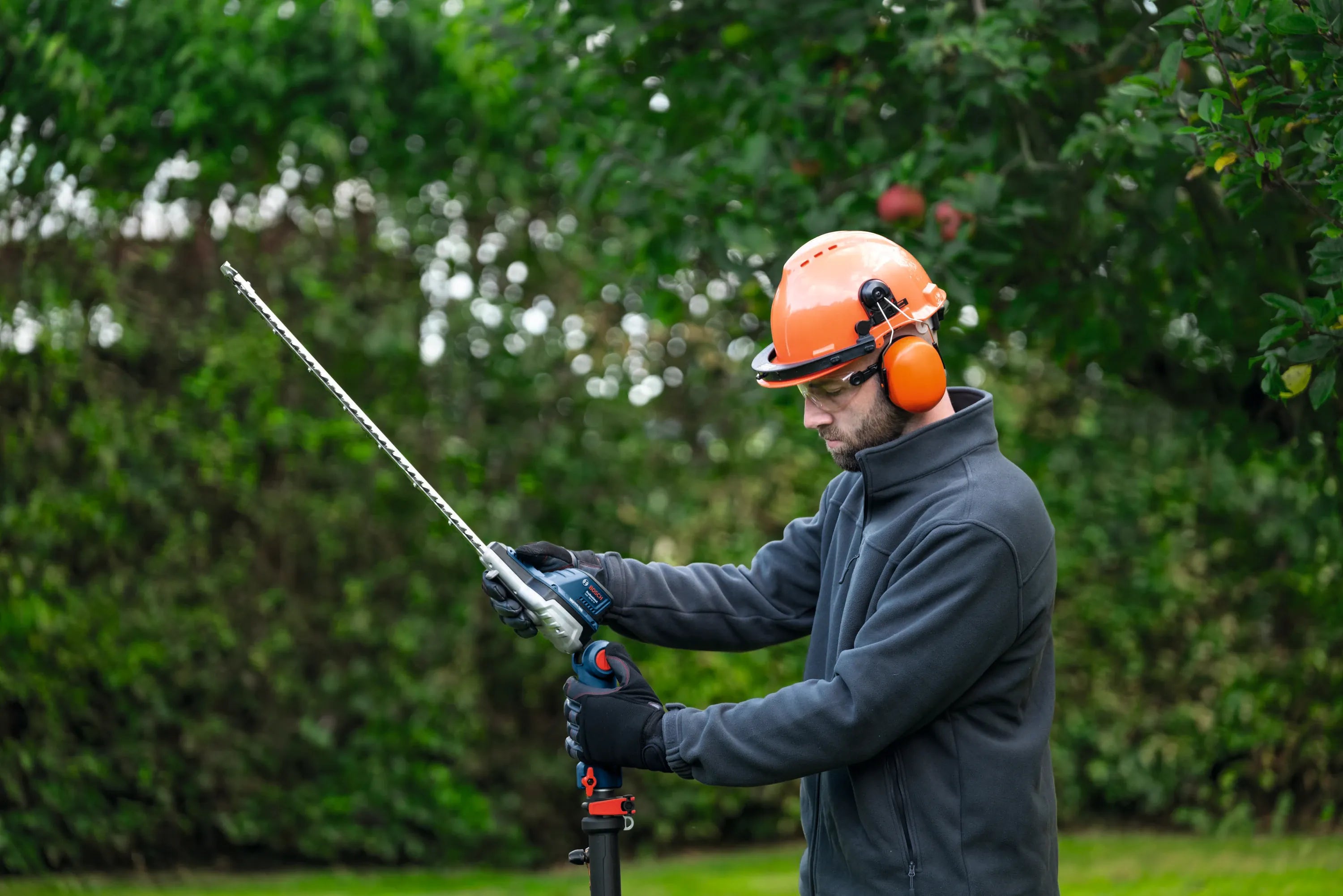 A side profile view of a property maintenance worker in an orange hard hat and earmuffs calibrating the multi-position articulating head joints on the cordless pole trimmer shaft before working on a high privacy screen.