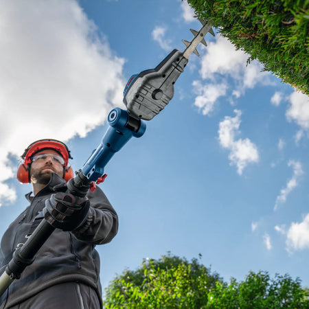 An under-head view of a gardener guiding the 20-inch diamond-ground laser-cut blade across a high decorative hedge corner. The operator uses both hands on the extended telescopic pole system to maintain straight, even cutting control under a clear blue sky.