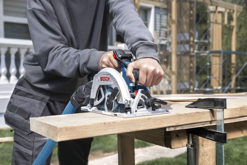 An outdoor construction scene showing a builder using the Bosch 18V brushless 7-1/4 inch circular saw to trim a thick structural framing lumber beam on an active outdoor job site, with uninstalled scaffolding structures visible in the background.