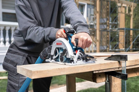 An outdoor construction scene showing a builder using the Bosch 18V brushless 7-1/4 inch circular saw to trim a thick structural framing lumber beam on an active outdoor job site, with uninstalled scaffolding structures visible in the background.