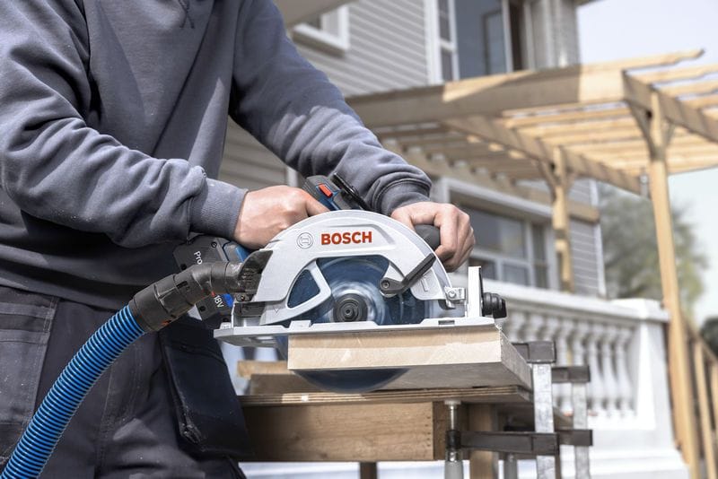 A builder uses the Bosch 18V brushless 7-1/4 inch circular saw to trim thick structural wood planks on an outdoor construction site. An attached blue dust extraction hose channels sawdust away from the cut line while a residential building framework stands in the background.