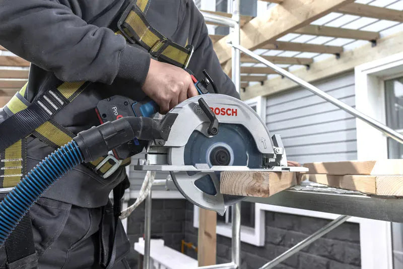 A construction worker wearing a safety harness operates the high-speed cordless framing circular saw on a metal scaffolding setup. The user cuts a piece of dimensional lumber while an integrated rotating dust chute extracts debris through a flexible vacuum system.