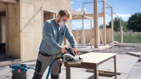 An outdoor job site view of a builder sizing wood materials on a work table with the Bosch GKS18V-68-2N circular saw. The high-capacity tool is paired with a portable dust extractor canister to maintain a clean, professional framing and installation environment.