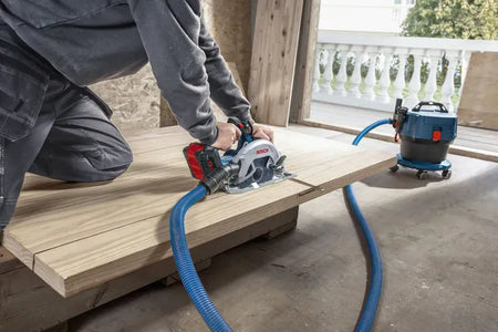 A carpenter operates the Bosch high-speed cordless circular saw to make a long crosscut along a wide wooden sheet on the floor. A flexible blue dust extraction hose connects the tool directly to a compact industrial wet-dry vacuum cleaner positioned in the background of the workshop.