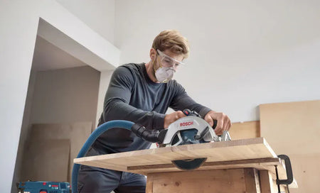 A low-angle view shows a craftsman wearing safety goggles and a dust mask using the Bosch cordless circular saw to cut a sheet of plywood mounted on a wooden sawhorse table. The integrated pivoting dust chute directs debris efficiently away from the cut line via a blue extraction vacuum hose system.