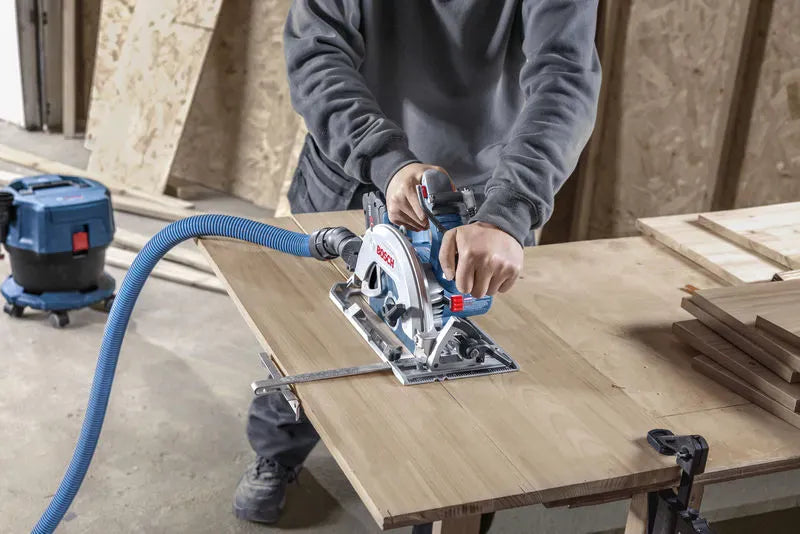 A close-up view of a carpenter making an accurate crosscut through a thick wood board clamped securely to a workbench table. The operator maintains firm control using both handles of the Bosch GKS18V-68-2N circular saw while an attached parallel fence guide tracks the straight edge.