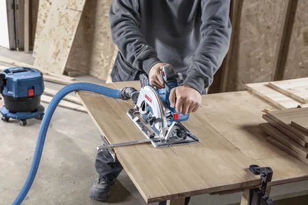 A close-up view of a carpenter making an accurate crosscut through a thick wood board clamped securely to a workbench table. The operator maintains firm control using both handles of the Bosch GKS18V-68-2N circular saw while an attached parallel fence guide tracks the straight edge.