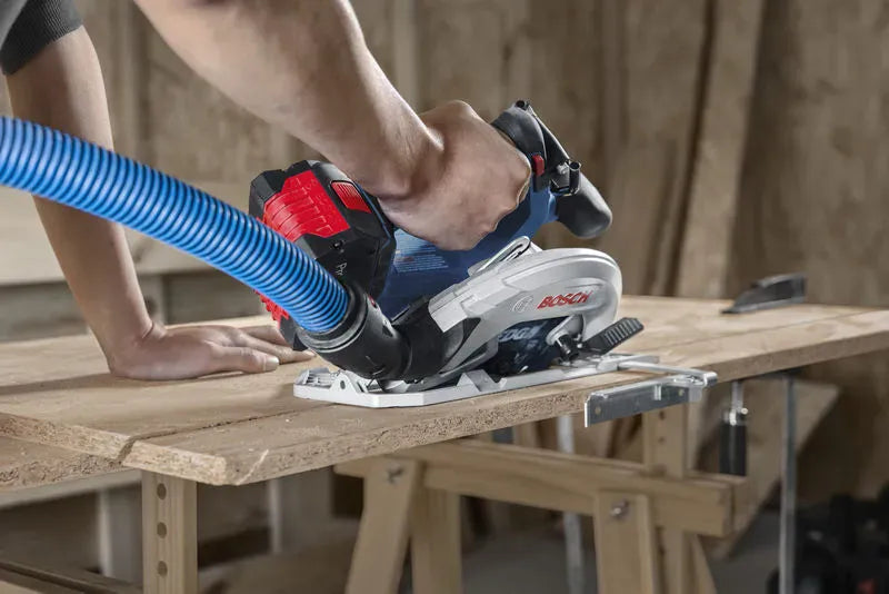 A detailed rear-angle view of a worker pushing the Bosch cordless wood cutting tool through a sheet of OSB composite board. The image underscores the tool's compact ergonomics and features a blue extraction hose connected securely to the rotating dust chute to maintain a clean workspace.