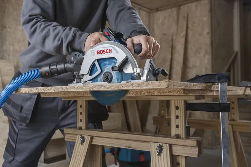 A front view of a craftsman using the high-torque circular saw to cut through a section of particle board resting on a pair of wooden sawhorses. The blue carbide-tipped wood blade can be seen extending cleanly through the bottom of the clamped workpiece.