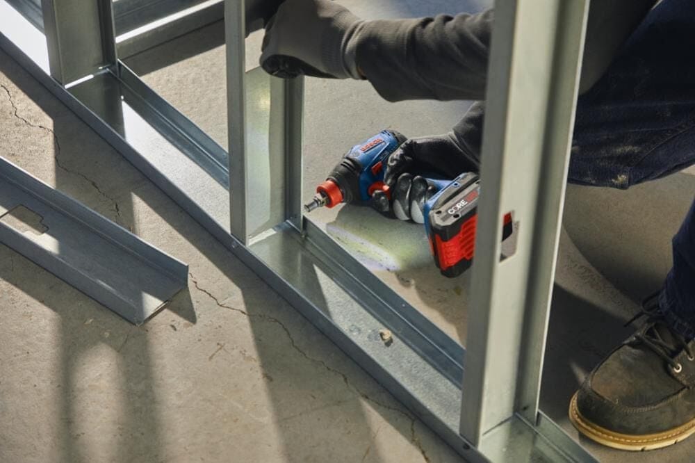 Worker fastening metal studs with power drill on concrete floor; gloves and boots worn in construction setting.