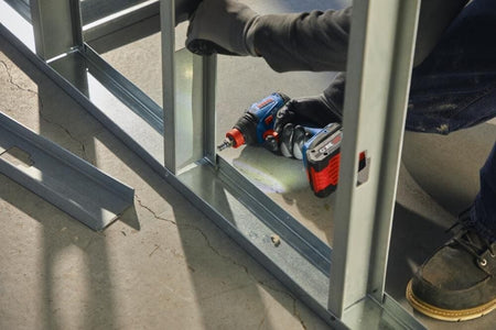 Worker fastening metal studs with power drill on concrete floor; gloves and boots worn in construction setting.