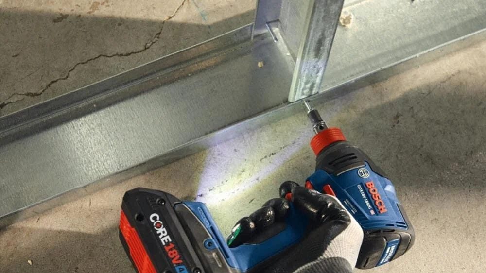 Bosch drill with CORE18V battery driving screw into metal stud frame on concrete floor; gloved hand visible.