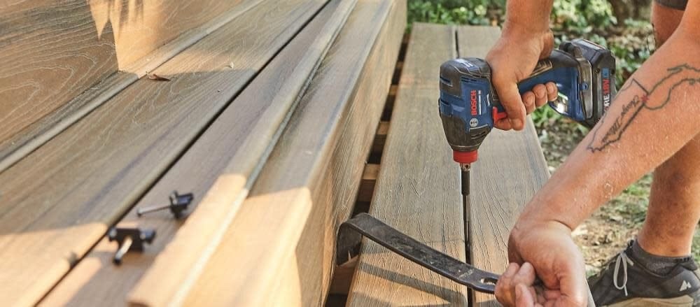 Person using Bosch cordless drill to drive screw into composite deck board; pry bar used for wood positioning.