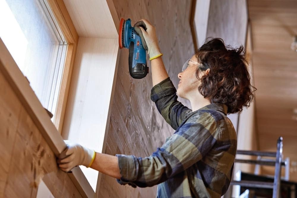 An action shot of a worker in safety glasses sanding an angled wooden window frame with the Bosch 12V sander.