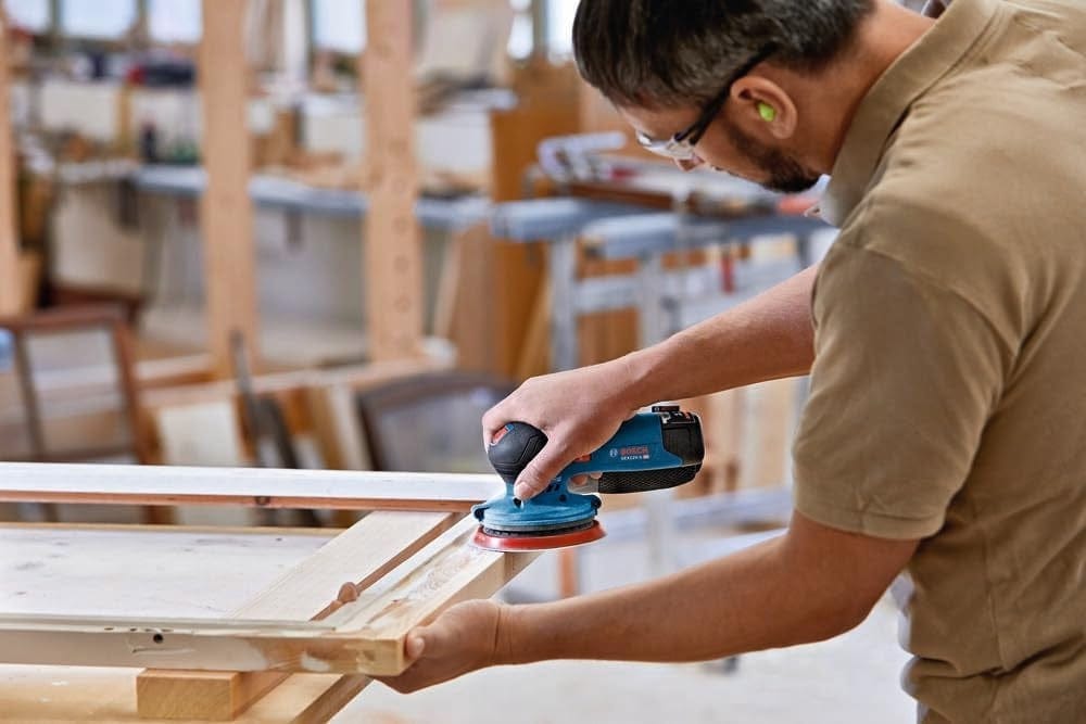 An action shot of a woodworker in a shop using the Bosch 12V sander to sand the profile of a wooden window frame.