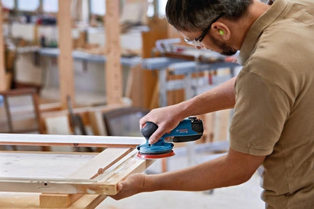 An action shot of a woodworker in a shop using the Bosch 12V sander to sand the profile of a wooden window frame.