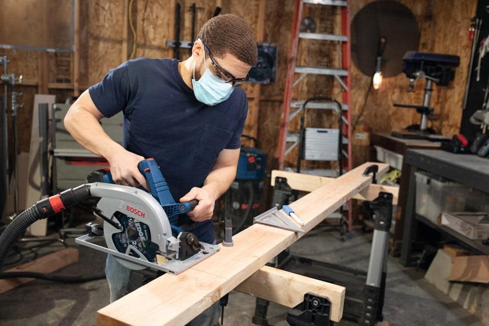 Person wearing safety gear using Bosch circular saw to cut wooden plank on sawhorses in a workshop setting.