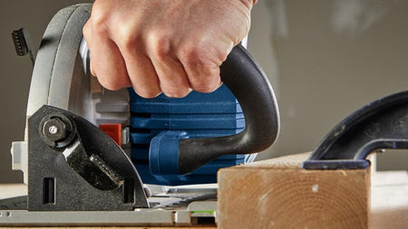 Person gripping Bosch circular saw while making a straight cut through wood secured with a clamp on workbench.