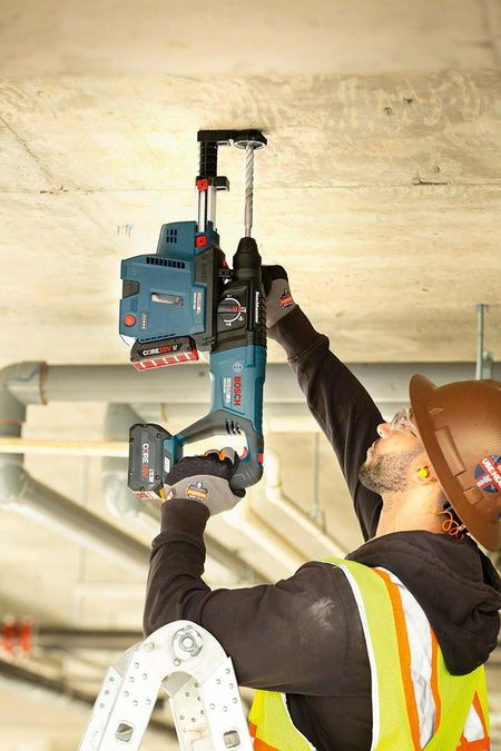 Worker drilling into concrete ceiling with Bosch cordless rotary hammer equipped with dust extraction system; standing on ladder in jobsite.