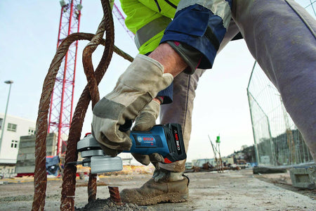 Construction worker in safety gear using Bosch cordless angle grinder to cut rusted rebar; sparks flying in active jobsite environment