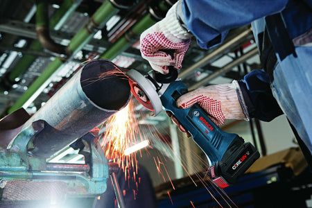 Bosch cordless angle grinder in use cutting metal pipe, producing sparks; tool held in vise on workbench in industrial workshop setting.