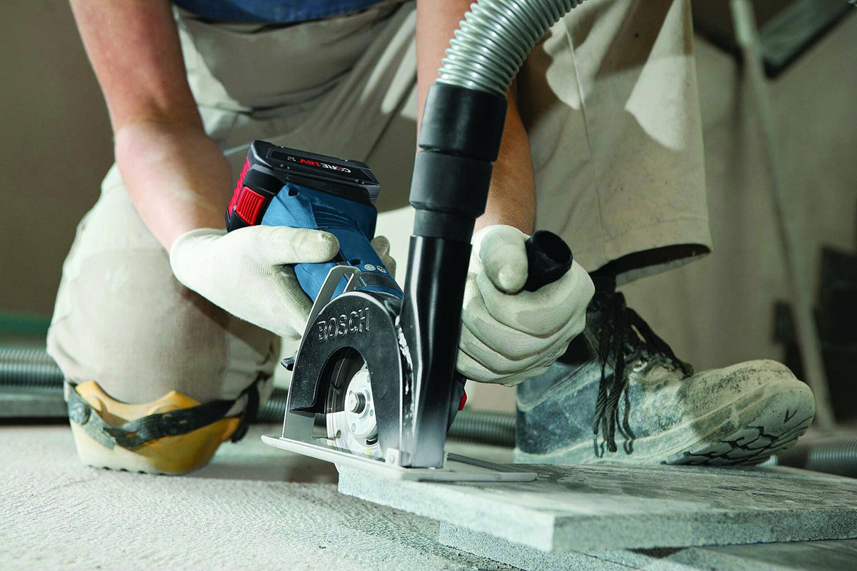 Worker using Bosch handheld grinder with vacuum attachment to cut or shape concrete, wearing gloves and boots at construction site.