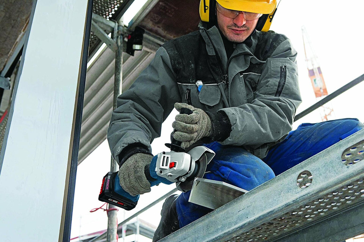 Construction worker wearing PPE operates Bosch cordless angle grinder on metal structure at jobsite with scaffolding and crane in background