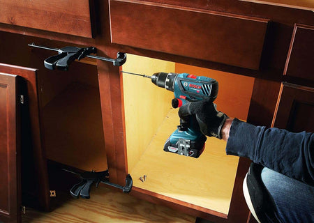 Person drilling into a wooden cabinet frame with a Bosch 18V cordless drill driver, showing controlled drilling, clamp‑secured alignment, and compact tool design ideal for cabinetry, woodworking, and interior installation projects.