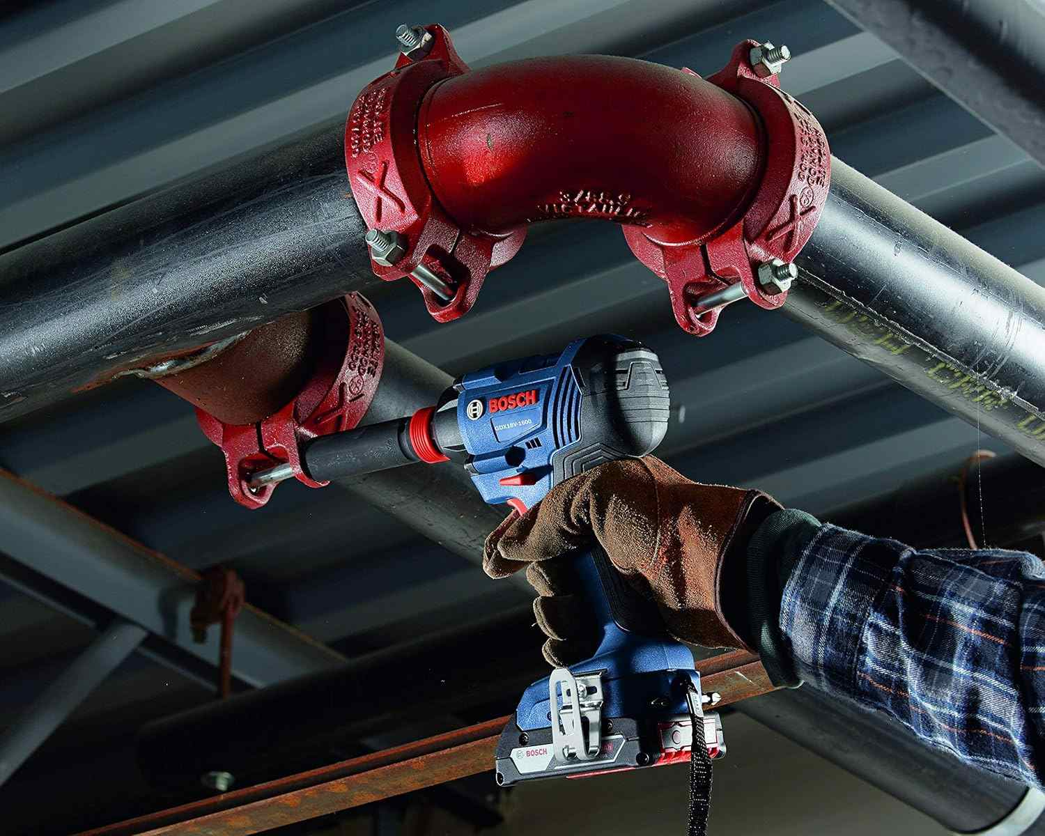 Worker using a Bosch 18V cordless impact driver to tighten bolts on a red pipe clamp assembly, highlighting high‑torque fastening capability, ergonomic grip, and dependable performance for plumbing, HVAC, and industrial maintenance work.