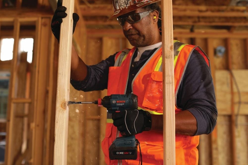 Construction worker drilling into a vertical wooden stud with a Bosch 18V cordless drill driver, emphasizing compact tough performance, active drilling power, and jobsite safety practices in framing and structural carpentry.