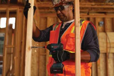 Construction worker drilling into a vertical wooden stud with a Bosch 18V cordless drill driver, emphasizing compact tough performance, active drilling power, and jobsite safety practices in framing and structural carpentry.