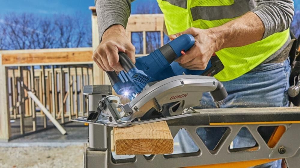 Worker in safety vest using Bosch circular saw on lumber; saw mounted on sawhorse with illuminated cutting area.
