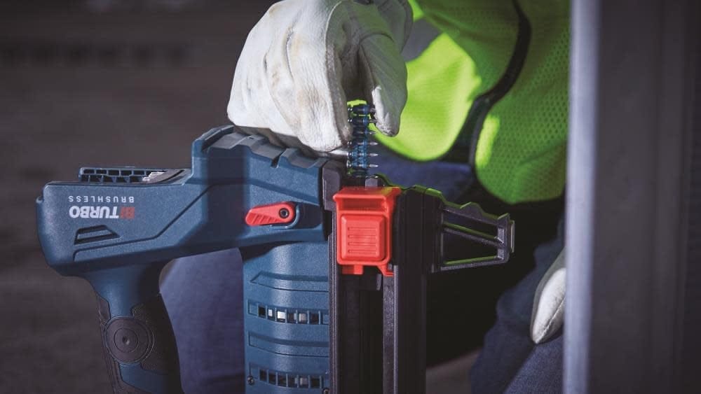 An action photograph of a worker's gloved hand inserting a strip of collated steel pin nails into the magazine of a Bosch GNB18V-12 cordless concrete nailer.