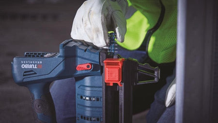 An action photograph of a worker's gloved hand inserting a strip of collated steel pin nails into the magazine of a Bosch GNB18V-12 cordless concrete nailer.
