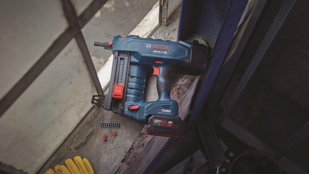 A wide action photograph showing the Bosch GNB18V-12 cordless concrete nailer resting on a concrete ledge next to a strip of collated steel nails and a pair of yellow work gloves.