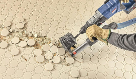 An action image demonstrating a worker using a Bosch rotary hammer and an angled flat scaling chisel with a dust attachment to strip up old hexagonal ceramic floor tiles and underlying hardened mortar beds quickly.