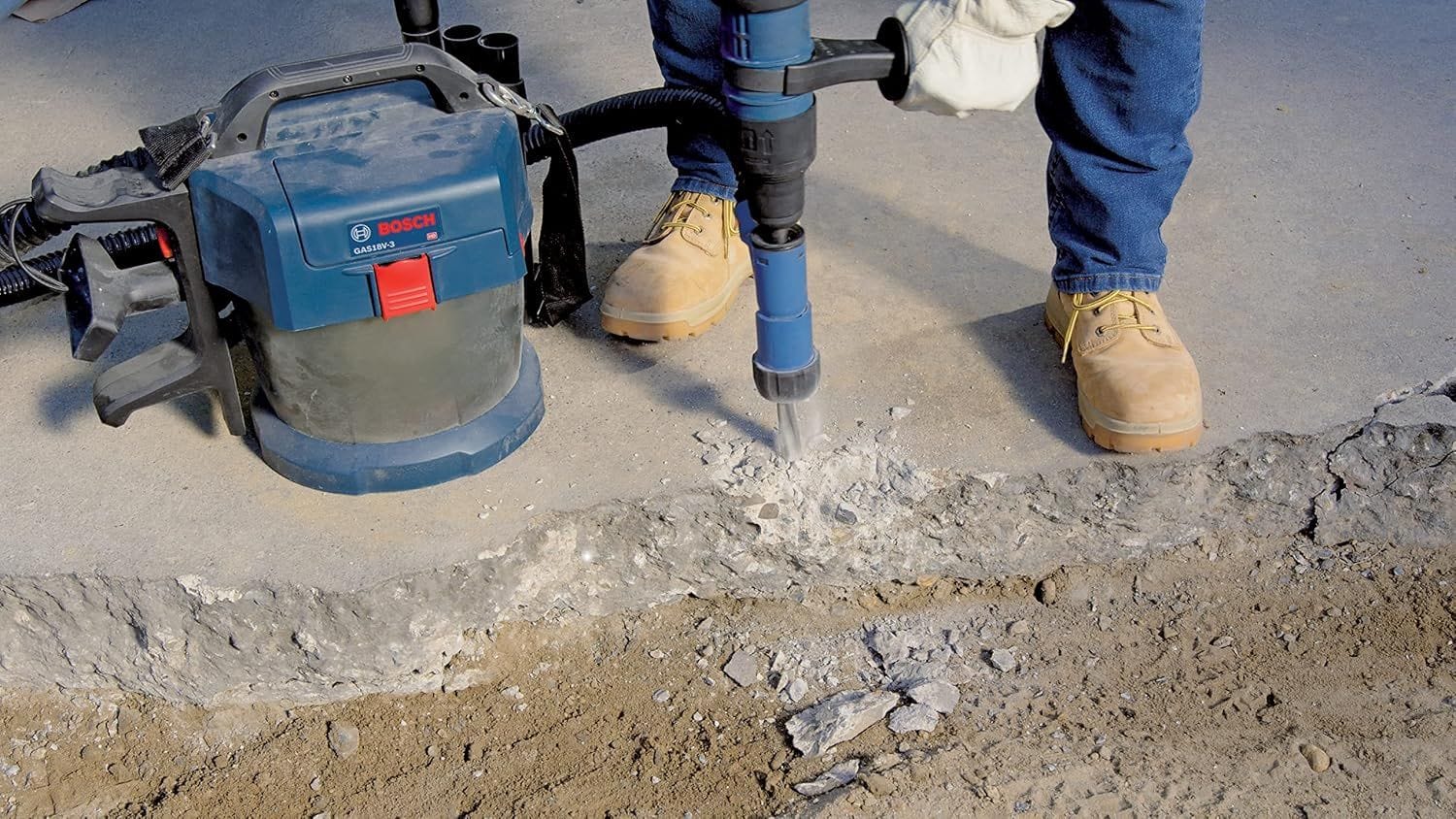 A low-angle jobsite photo of a worker wearing leather gloves and boots operating a Bosch cordless demolition tool to break up a concrete foundation edge next to a portable Bosch cordless dust extractor vacuum.