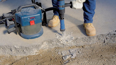 A low-angle jobsite photo of a worker wearing leather gloves and boots operating a Bosch cordless demolition tool to break up a concrete foundation edge next to a portable Bosch cordless dust extractor vacuum.