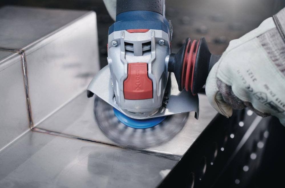Person using angle grinder with blue abrasive disc to polish metal surface; protective gloves worn in workshop setting.
