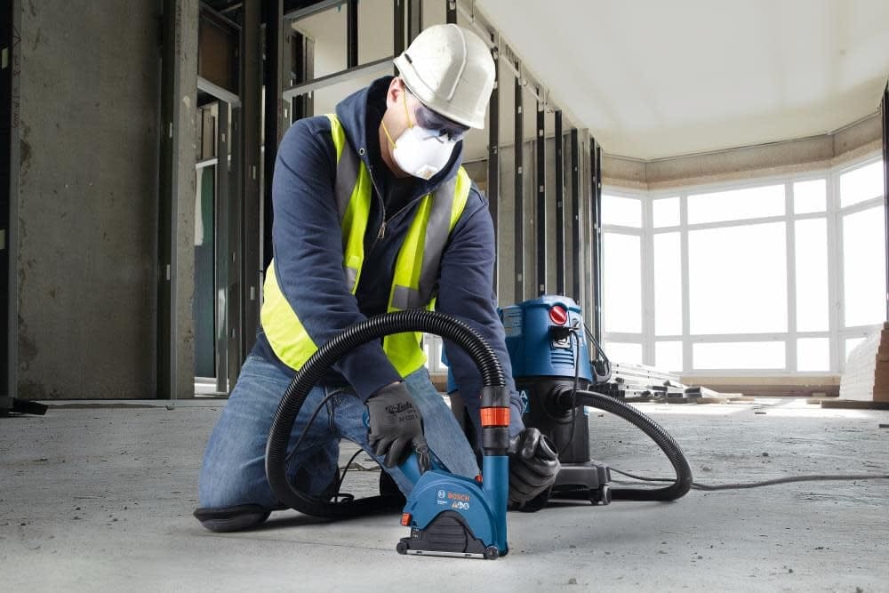 Worker using Bosch tool on concrete wall with HEPA vacuum system; industrial pipes and valves overhead.