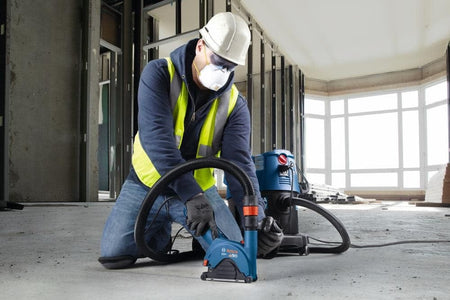 Worker using Bosch tool on concrete wall with HEPA vacuum system; industrial pipes and valves overhead.