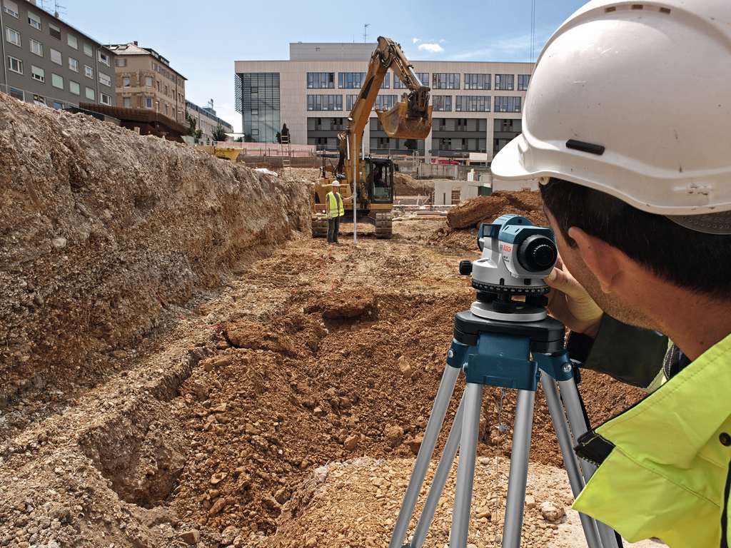 Surveyor operating tripod-mounted instrument near excavator and urban buildings during active excavation work.
