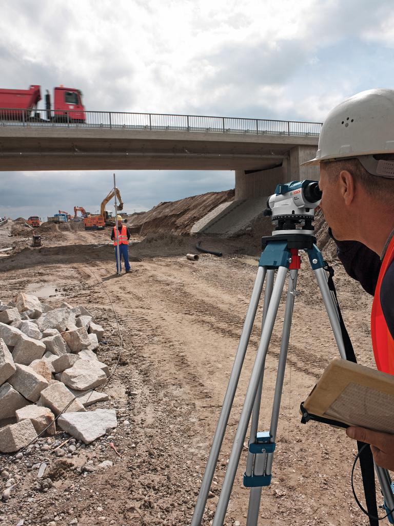 Surveyor using tripod-mounted optical level on construction site; bridge, truck, and excavator visible in background.