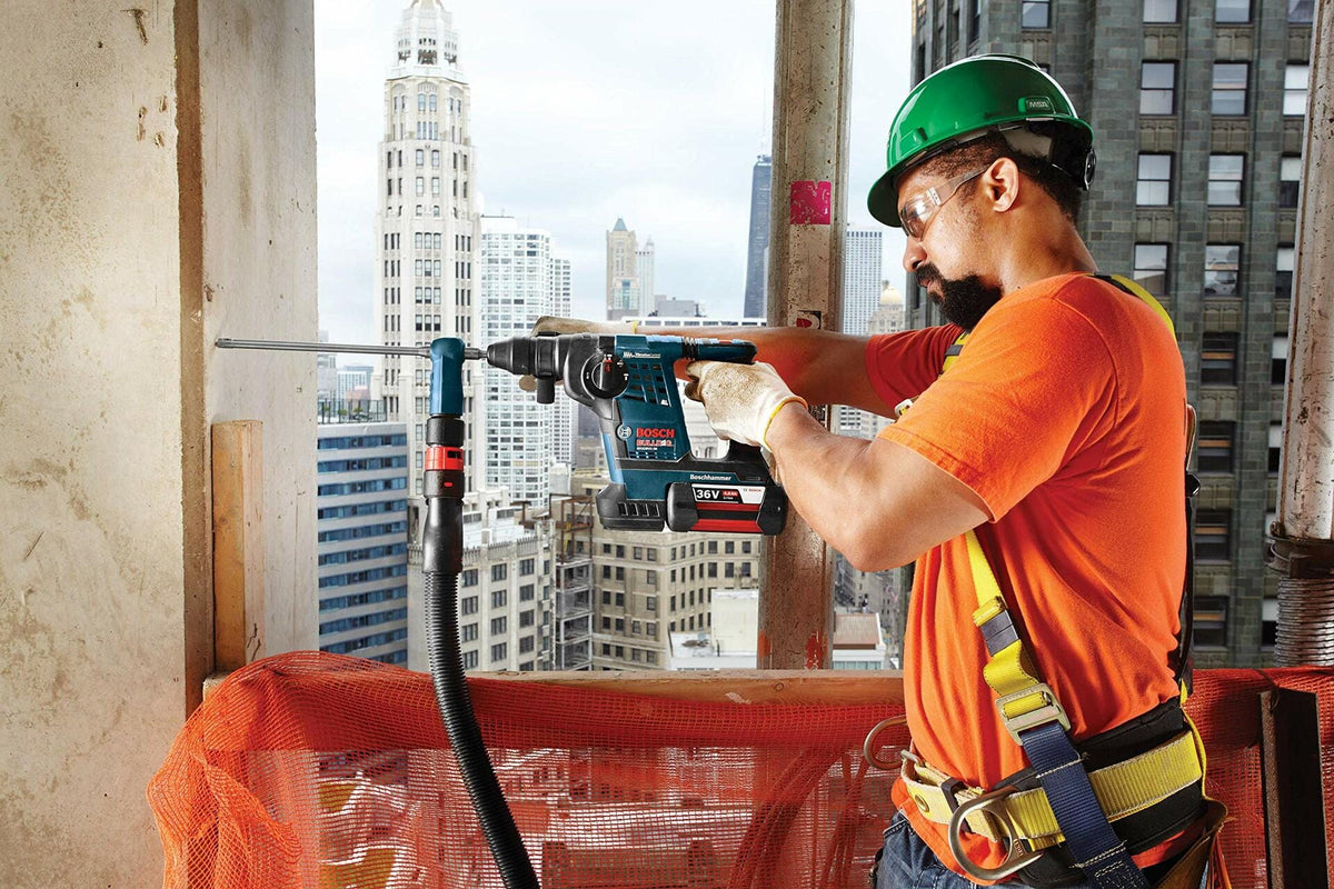 Construction worker using Bosch rotary hammer drill with dust extractor on high-rise site; city skyline visible in background.