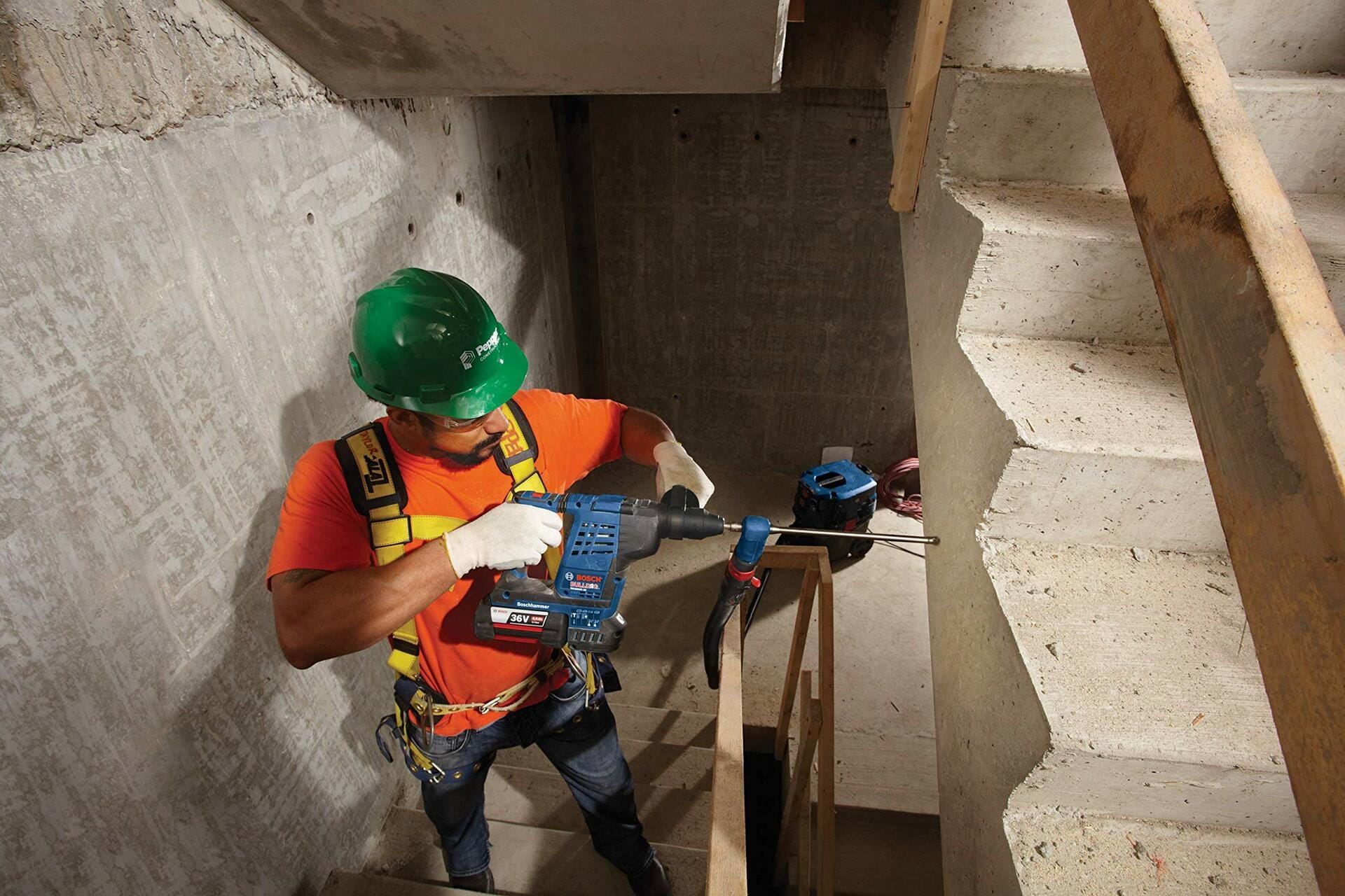 Worker in harness and PPE using rotary hammer drill on concrete wall in stairwell construction zone; standing on temporary platform.