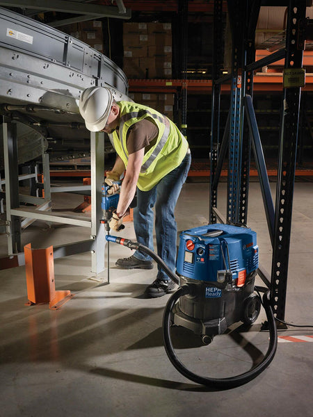 Worker using Bosch power tool with HEPA vacuum to drill or fasten metal structure in warehouse; wearing full safety gear.