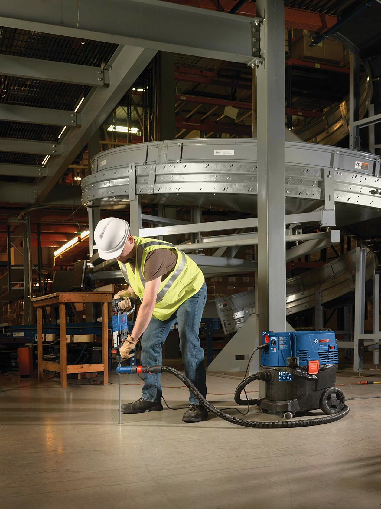 Person in hard hat and safety vest using Bosch power tool with vacuum system to drill into concrete floor in industrial facility