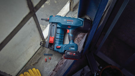 Bosch GNB18V-12 cordless concrete nailer placed on a concrete ledge with red plastic components and a strip of collated nails nearby. Tool features a Turbo battery and is shown in a workshop setting, ready for use.